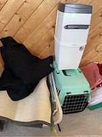 Small plastic cat carry cage, white plastic litter container with lid, black fabric cage cover, and other accessories stacked on a beige mat against wood wall.