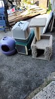 Photo showing set of four cat care items: a plastic cat carrier, two litter boxes (purple dome and green tray), and beige carpeted scratching post with platform and cubby hole base.