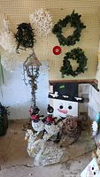 Wide view of two green wreaths hanging on pegboard, white decorative snowflakes, snowman figure, and lighted wire birds and swan figures on the floor