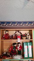 Photo showing wooden shelf with two stuffed holiday figures in festive clothes, pinecone owl ornaments, small wood schoolhouse, wreath, sleigh, and some snowflake ornaments placed on the shelves.