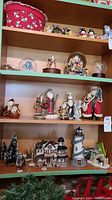 Multiple Christmas figurines including Santa Claus figures, snow globes, the Harbortview Inn village house, a lighthouse, and Christmas trees arranged on wood shelves.