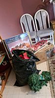 Photo showing boxed 16-piece electric lighted Dickens ceramic village, black garbage bag likely containing Christmas decorations, pine garland bundle, and cardboard box for tree or decorations.
