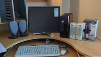 View of Dell desktop tower, 19-inch Dell monitor, two sets of computer speakers including black and white sets, Blue Yeti Nano microphone box, HP wired keyboard, and wired mouse on wooden desk.