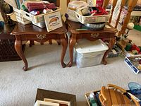 Two wooden end tables with drawers, cabriole legs, and brass drawer pulls shown with various household items on top and around them.