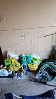 Display of gardening supplies including hoses, hose reel, bags of soil, watering cans, sprayers, hand tools, and tomato cage against a concrete wall