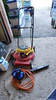 Photo of red Noma electric lawnmower with foldable handle and yellow extension cord, along with blue Sun Joe leaf blower with orange cord placed in front.