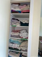 Full view of linen cupboard shelves showing stacks of sheets, duvet covers, pillowcases, runners, towels, and blankets in varied colors and patterns.