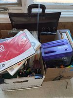 Photo showing cardboard boxes filled with office supplies including lined paper packs, binder clips, pens, and a purple Brinks mini shredder.