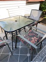 Photo showing square glass top patio table with black metal frame and four matching metal mesh chairs with floral cushions arranged on outdoor rug.