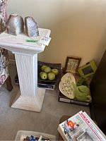 View of off-white fluted pedestal, geode bookends on top, faux green apples in wire basket, decorative plates, picture frame, and marble apple.