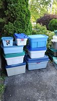 Stack of nine Rubbermaid plastic storage bins in various sizes and lid colors (blue, green). Bins are arranged outdoors on pavement near greenery.