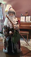 Full front view of freestanding Santa Claus figurine showing detailed face, curly white beard, and red robe with fur trim. Holding a long wooden staff and green sack with pine cone detail.