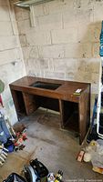 Wooden sewing table positioned against a concrete wall, showing inset opening on tabletop and overall structure.
