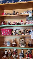 Wide view of multiple shelves showing various Christmas themed snowglobes, figurines including Santa Claus, snowmen, angels, nativity scene parts, and a red fabric box with snowman pattern