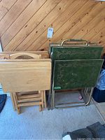 Two sets of TV trays and stands, pine wood set on the left and vintage green metal set on the right, standing against a wooden wall.