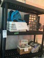 Side view of black resin utility shelf with varied contents on middle and top shelves including unpacked small white trays with note, blue bag and black drawer unit.