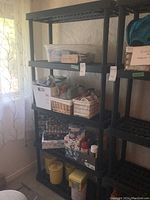Photo of black resin shelving unit filled with various household items including plastic containers, baskets, and packaged goods near a window with curtain.