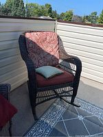 Brown resin wicker-like rocking chair with red and white patterned cushions and a plain green pillow on an outdoor deck.