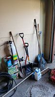 Photo showing 3 shovels, 2 rakes, broom, spray applicator, black coiled hose, and other garden items leaning against a wall.