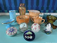 Mixed lot of glass items on green table, including vintage peach Fire King kitchenware, glass paperweights, various colored glass vases and goblets.