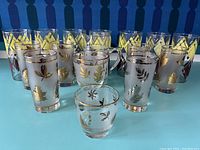 Full view of 14 glass pieces showing tall yellow leaf highball glasses, frosted gold leaf lowball glasses, small shot glass, pitcher, and goblet, in front of a blue patterned background