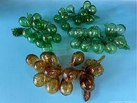 Four glass grape clusters on blue background, three green and one amber. One green cluster has a broken detached leaf visible.