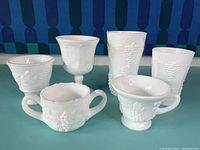 Front view of 5 different milk glass cups, all with grape and leaf embossed pattern.