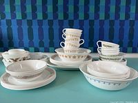 Full lot of mixed vintage Pyrex and Corelle patterned dinnerware including cups, plates, bowls, and small serving dishes arranged on turquoise surface against blue patterned background.