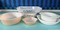 Seven vintage glass baking dishes of various shapes and designs on blue table with patterned backdrop