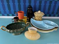 Mixed lot of colorful vintage Tupperware cups stacked behind green decorative bowl with gold handles, 4 stacked divided ceramic plates in pastel blue, white, yellow and brown, yellow canister with textured lid, and amber jar in background. Items arranged on blue surface with blue patterned backdrop.