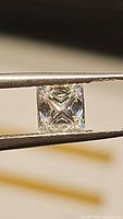 Close-up image of the princess cut moissanite gemstone held by tweezers showing cut and clarity.