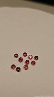 Nine small round cut fancy colored sapphires in red, pink, orange tones on a neutral background, showing their size and clarity.