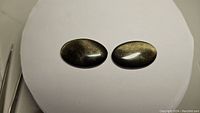 Pair of oval golden sheen obsidian cabochons placed on a white surface showing their golden reflective sheen.
