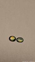 Pair of oval cabochon cut natural multicolored sapphires, green-yellow-blue hues, resting on beige background.