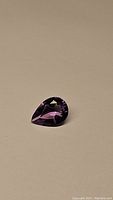 Close-up photo of pear shaped purple amethyst gemstone showing faceting and clarity against plain beige background.