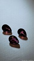 Three natural fancy colored pear-shaped garnet gemstones displayed on a white surface showing rich hot pink to reddish-orange colors.