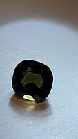 Images showing the natural olive green cushion cut tourmaline gemstone resting on a surface under lighting to highlight the color and facets.