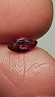 Close-up photo of the marquis cut ruby gemstone held between fingers, showing its deep red color with hot pink undertones and clarity.