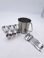 Photo showing three vintage metal ice cream scoops with spring mechanisms, a stainless steel measuring cup with interior volume markings and handle, and six forks laid out.