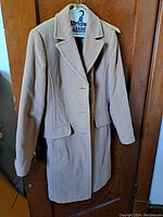 Full view of the Lebek tan wool and cashmere overcoat hanging on a door, showing the front buttons, lapels, and side pockets.