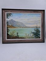 Front view of oil painting on panel showing coastal mountain, water, and vegetation scene in wood frame with decorative nail heads.