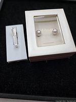 View of the pair of pearl earrings in original Misako box alongside the silver-tone arrow tie clip with a small pearl detail, both displayed on white holders against a black background.