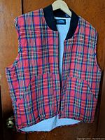 Full front view of Dutch Harbor Gear sleeveless red tartan vest with fleece lining hung on wood door