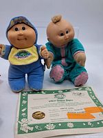 Two vintage Cabbage Patch Kids dolls with official adoption papers placed in front of them on a white background.