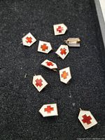 Top view of eight small shield-shaped pins with white enamel and red cross design scattered on dark cloth surface.