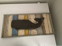 Framed art piece with a carved or raised grey whale on multi-colored distressed wood slats.
