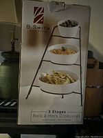 Photo of box with image of B. Smith 3 Tier Server Tray Set showing three white porcelain bowls stacked on a collapsible black metal stand.