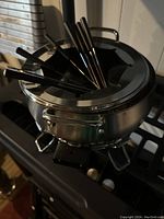 Electric Cuisinart fondue pot sitting on a stove, showing the brushed stainless steel pot with lid that holds six black-handled fondue forks inserted into slots.