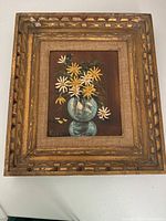 Photo of framed oil painting showing yellow and white flowers in a blue vase on a brown background.