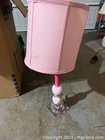 Tall pink lamp with fabric shade shown on concrete floor next to cardboard box, light pink color with clear glass base and pink accents, square glass base visible.